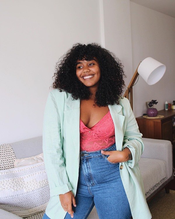 Mulher jovem e negra, com cabelos cacheados médio, posando para foto na sala de casa. Ela usa um blazer verde água, um top de renda rosa e calça jeans.