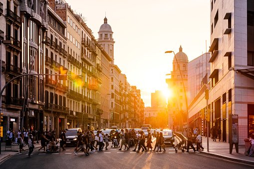 Pessoas cruzando uma faixa de pedestres em rua movimentada na Espanha. Em ambos os lados é possível ver prédios e ao fundo, o sol do final da tarde - Metrópoles