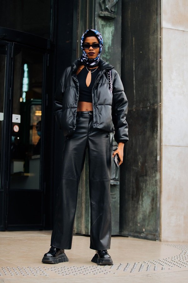 Mulher jovem e negra, provavelmente uma modelo, durante a Semana de Moda de Paris, na França. Ela usa um top curto, uma jaqueta puffer e uma calça de couro, todas as peças são pretas.