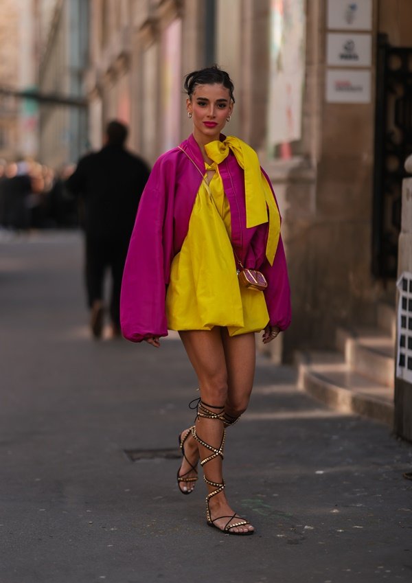 Mulher jovem e branca, com cabelo castanho amarrado, durante a Semana de Moda de Paris, na França. Ela usa um vestido amarelo bufante com casaco lilás e uma sandália estilo gladiadora.
