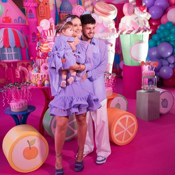 A influenciadora Virgínia ao lado do seu namorado Zé Felipe no aniversário da sua filha de um ano. Todos usam roupas lilás e a decoração do aniversário é rosa, com carrinhos de criança.