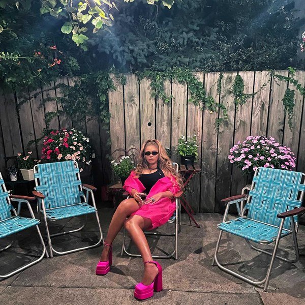 A cantora Beyoncé sentada em uma cadeira de praia, porém no quintal de casa. Ela usa um vestido preto, um casaco rosa e uma sandália de salto plataforma rosa