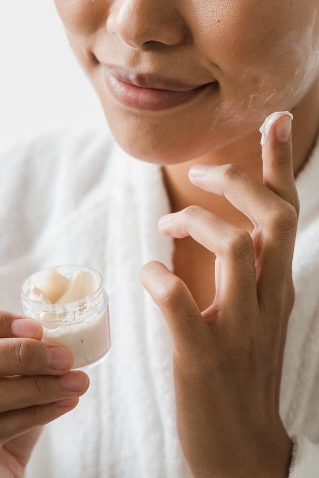 Meio rosto de uma mulher com meio sorriso passando creme no rosto com uma das mãos - Metrópoles