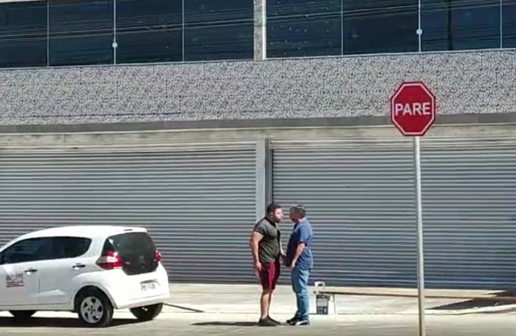 Homens se encarando ao lado de carro branco e de placa de Pare na rua