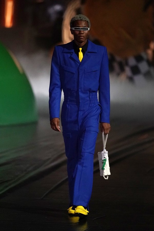 Modelo jovem e negro, com cabelo curto crespo, desfilando na passarela com roupas da marca Louis Vuitton. Usa um macacão azul royal, camisa preta e gravata amarela, além de uma bolsa de couro branca