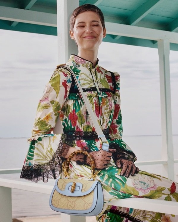 Mulher branca e jovem, com cabelos castanhos curtos, posando para foto em um barco. Ela usa um vestido estampado e uma bolsa azul clara com alça de bambu da marca Gucci