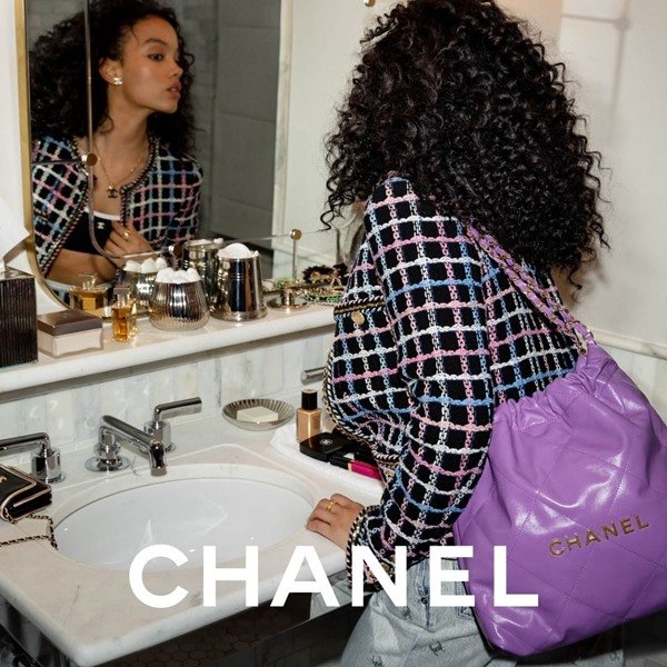 Mulher negra e jovem, com cabelos castanhos e cacheados, posando para foto em frente ao espelho do banheiro. Ela usa um top preto curto, calça jeans e um blazer de tweet preto e branco. Ela segura uma bolsa estilo saco lilás da Chanel.