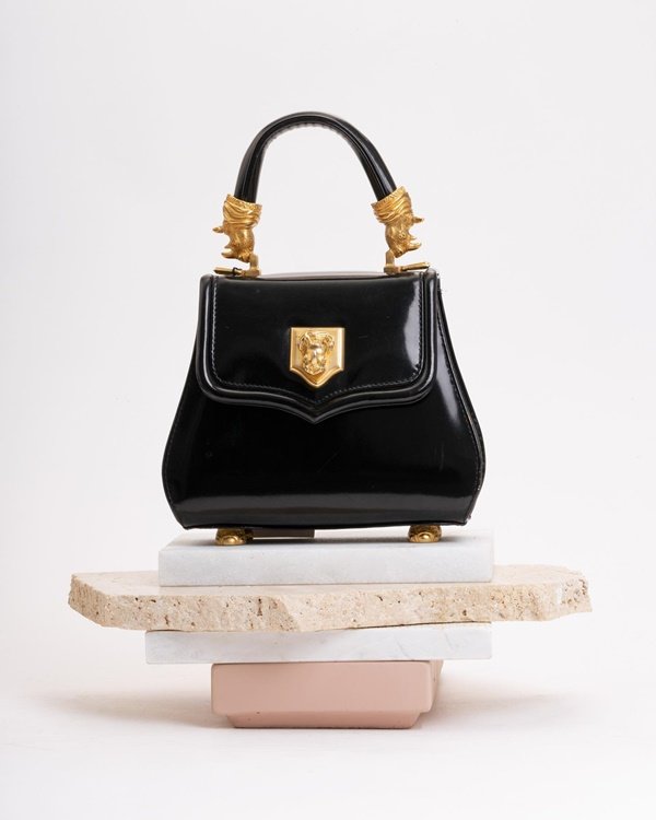 Bolsa preta e pequena de couro, com detalhes dourados, da marca Vicenza. O item está em cima de tábuas de mármore rosa e cinza e o fundo da foto é branco.