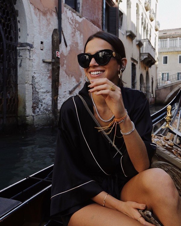 A atriz Bruna Marquezine, uma mulher branca, jovem e com cabelo castanho curto, posando para foto em um barco pelos canais de Veneza, na Itália. Ela usa um kimono preto com costuras brancas, óculos escuros e acessórios como colar, anéis e pulseira em prata.