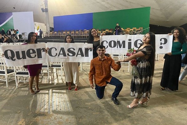 Foto de um homem ajoelhando pedido a mão de uma mulher em casamento com um cartaz ao fundo