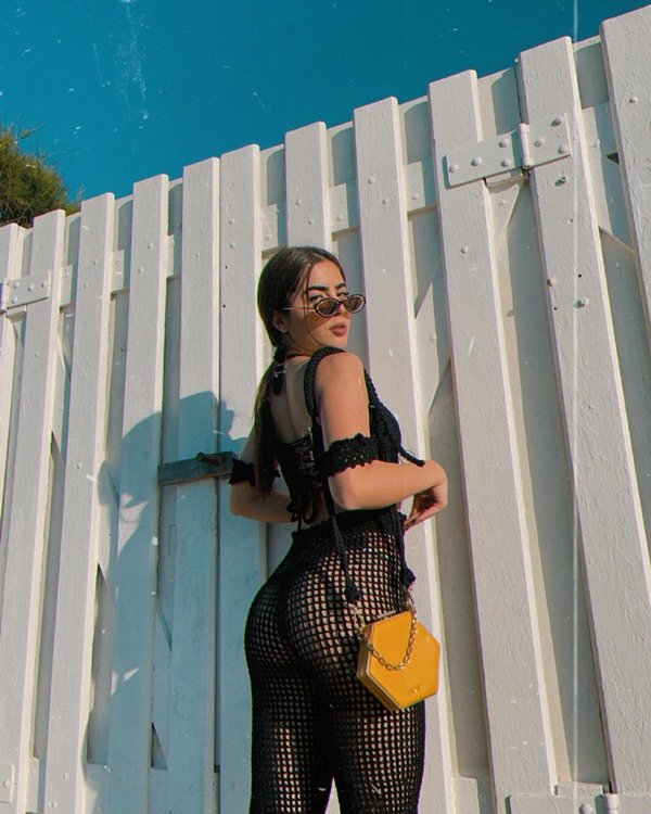 A influencer e ex-BBB Jade Picon posando para foto em uma cerca de madeira branca. Ela usa um top e uma calça de crochê pretos, óculos escuros e uma bolsa transpassada amarela
