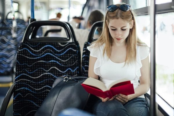 Mulher branca e jovem, com cabelo liso e longo, segurando um livro. Ela está sentada no banco do ônibus e usa uma blusa brancade babado e óculos escuros na cabeça.