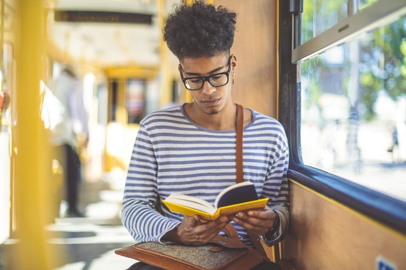 Homem jovem e negro, com cabelo cacheado, lendo um livro enquando anda de ônibus. Ele usa uma blusa de manda longa listrada nas cores brancas e azul e, no seu colo, leva uma pasta de couro marrom