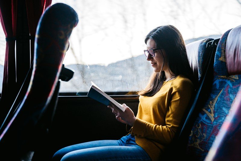Mulher branca e jovem, com cabelo liso e longo, segurando um livro. Ela está sentada no banco do ônibus e usa óculos de grau, calça jeans e uma blusa de manga longa amarela