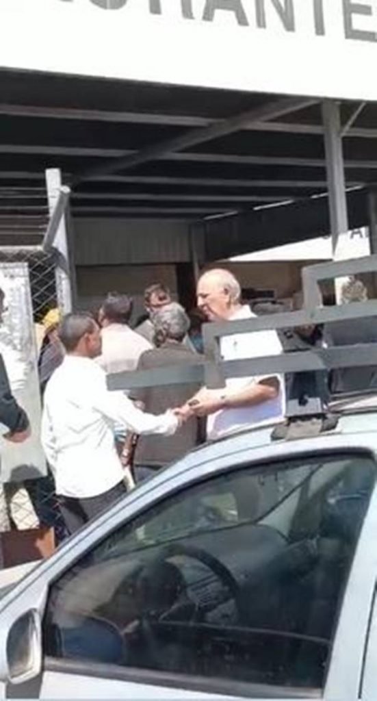 Imagem colorida de pessoas na fila e homem de camisa branco cumprimentando cidadãos