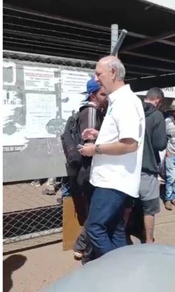 Imagem colorida de pessoas na fila e homem de camisa branco cumprimentando cidadãos