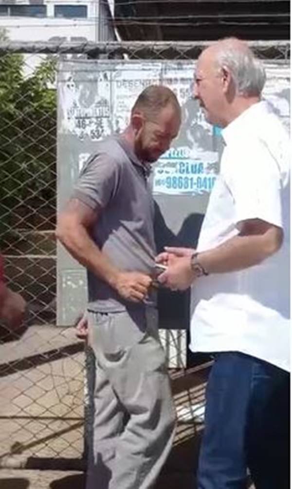 Imagem colorida de pessoas na fila e homem de camisa branco cumprimentando cidadãos