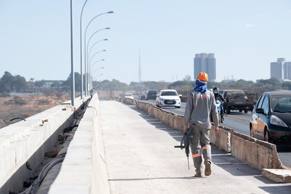 Foto de um operário andando por uma passarela de concreto com carros passando na pista ao lado