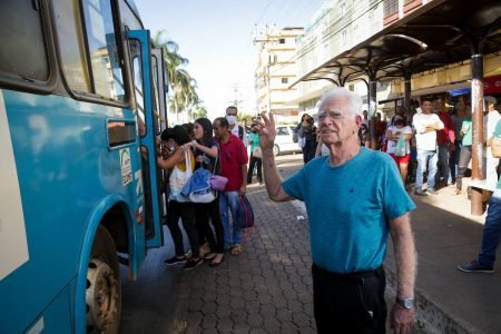 Homem idoso de blusa azul chama homem em parada de ônibus