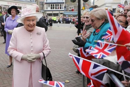 Foto colorida. Mulher idosa com roupa rosa claro lado de pessoas com bandeiras da Grã-Bretanha