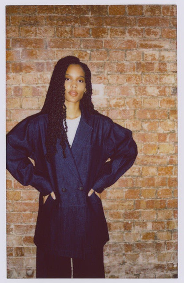Foto estilo polaroid que divulga a coleção de roupas da marca Asantii. Na imagem é possível ver uma modelo negra e jovem, de cabelos longos trançados, com uma camiseta branca e um casaco azul por cima