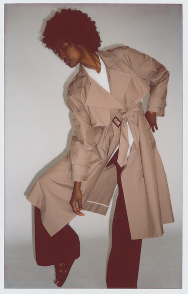 Foto estilo polaroid que divulga a coleção de roupas da marca Asantii. Na imagem é possível ver uma modelo negra e jovem, de cabelos cacheados curtos, com uma camiseta branca e um casaco bege estilo sobretudo por cima