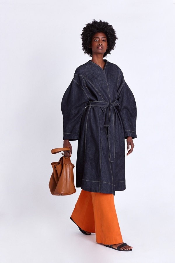 Foto estilo lookbook da marca Asantii, quando a modelo posa com a roupa em um fundo infinito branco. Na imagem é possível ver uma modelo negra e jovem de cabelo comprido trançado usando uma calça laranja e um casaco azul
