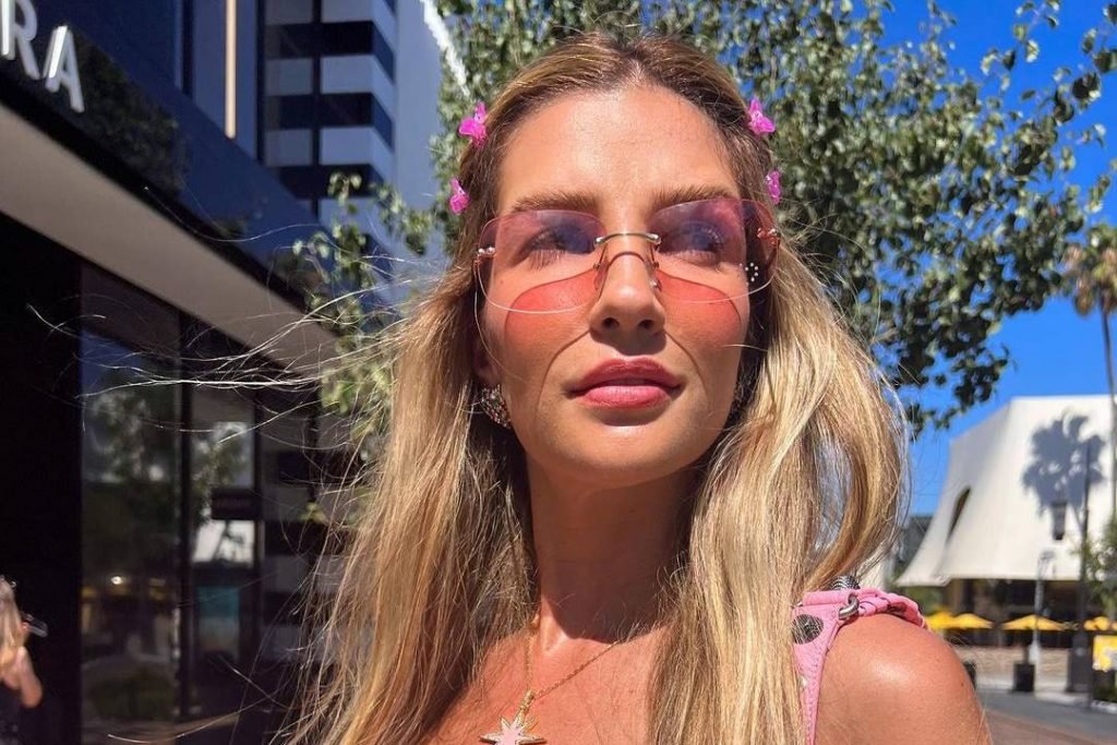 Aline Gotschalg com cabelo loiro, óculos com armação rosa em uma avenida - Metrópoles