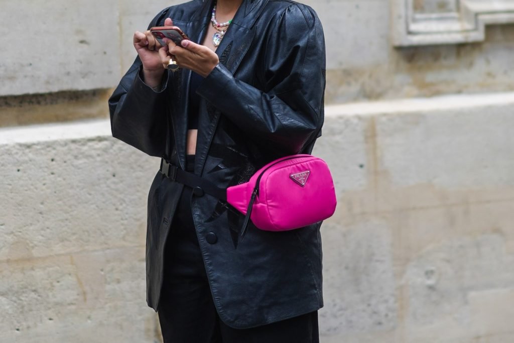 Mulher com roupa toda preta, mexendo no celular na rua. Ela tem amarrada na cintura uma pochete rosa da marca Prada