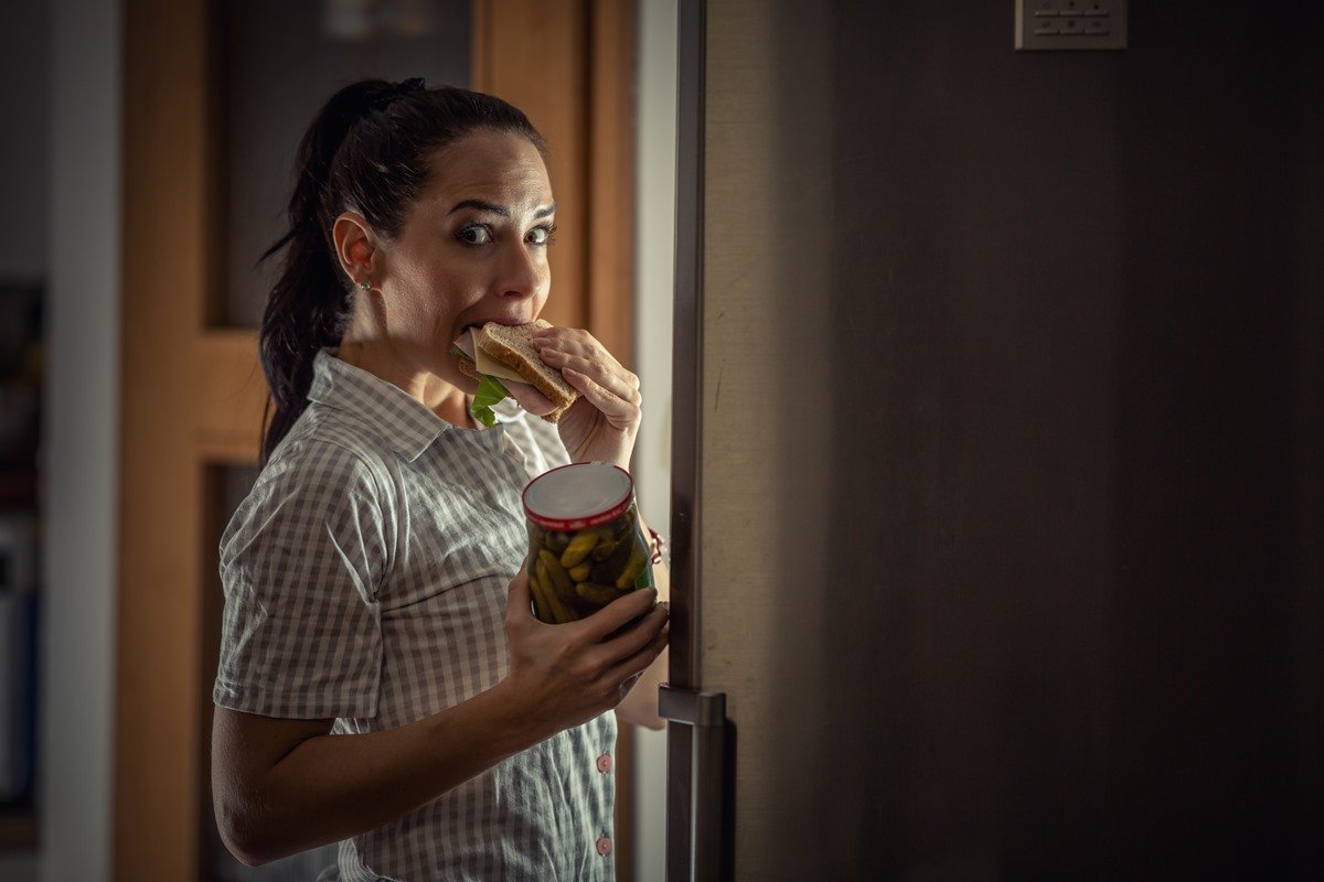 Woman raiding fridge at night bites into a sandwich holding pickled cucumbers with guilty facial expression.