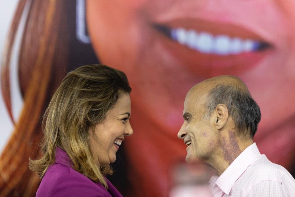 Mulher loira com blazer roxo virada de lado e sorrindo. Ela olha para um homem velho e careca, que também sorri para ela