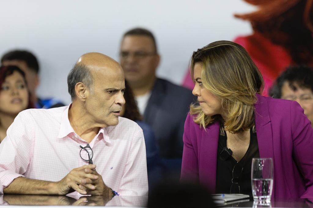Homem careca sentado com cara séria e rosto par o lado falando com mulher loira com blazer roxo