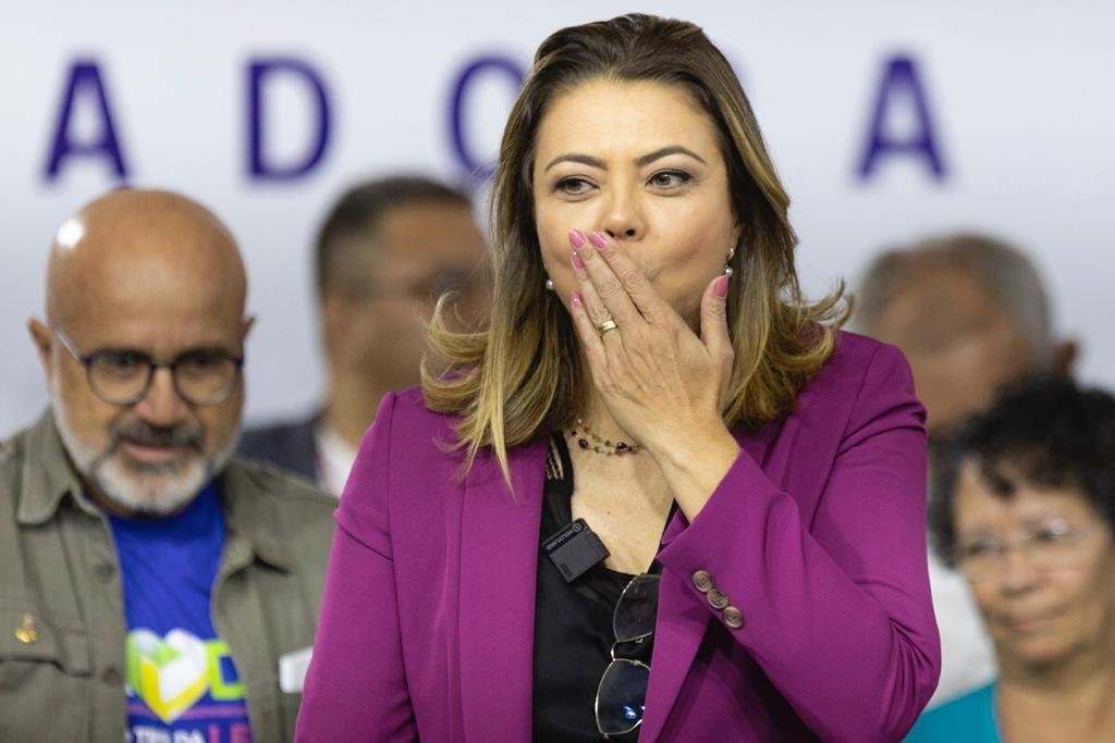 Mulher loira com blusa preta e blazer roxo com a mão na boca mandando beijo