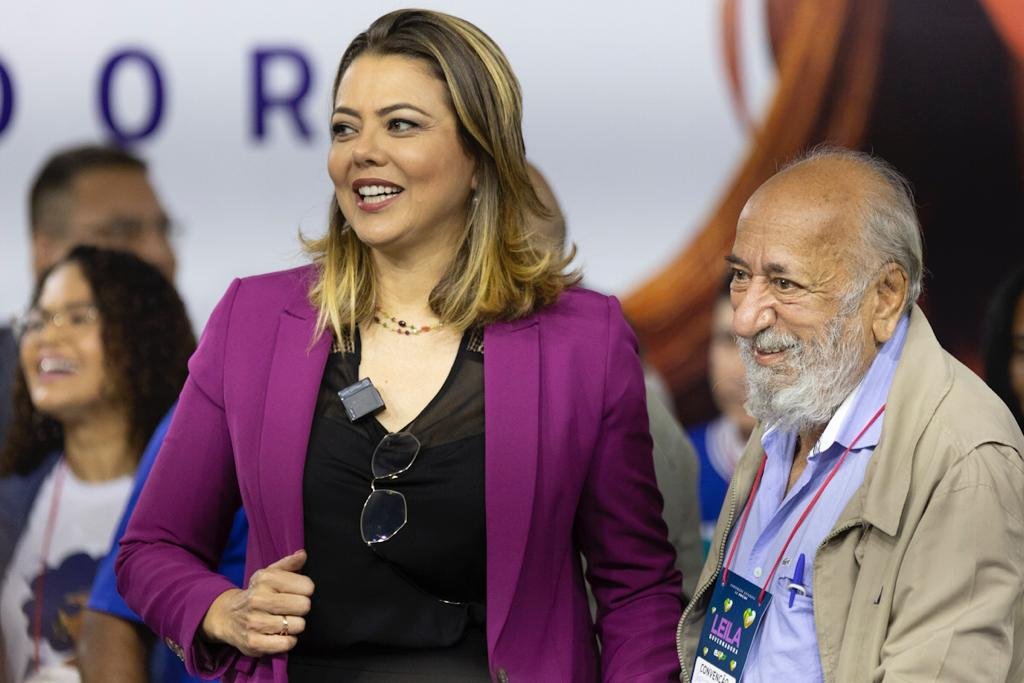 Mulher loira com blusa preta e blazer roxo ao lado de idoso com casaco bege