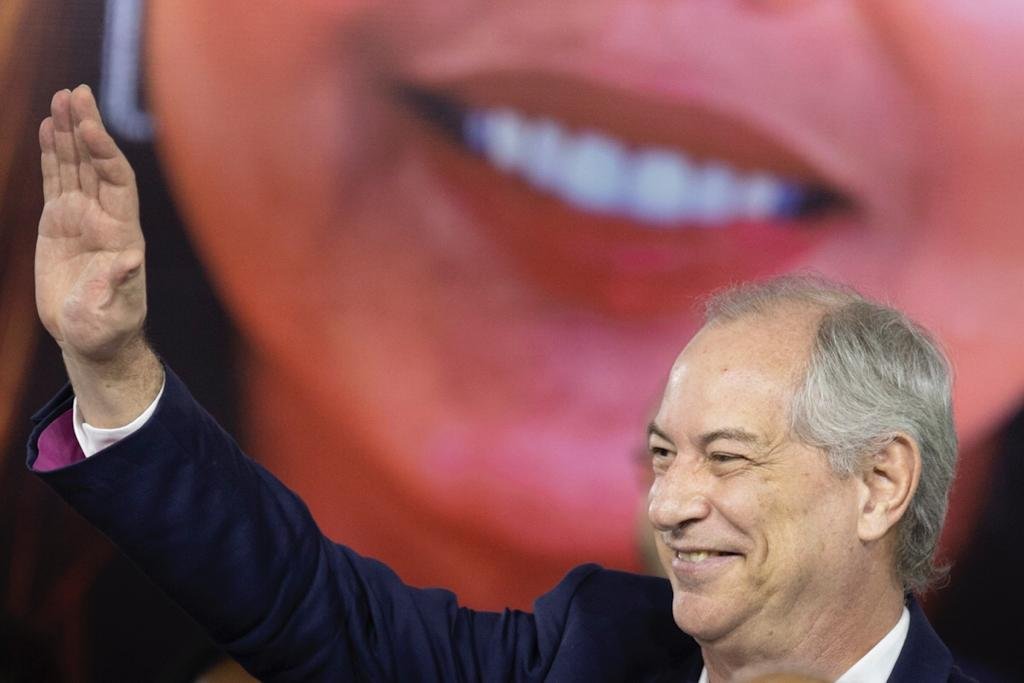 Homem branco, calvo e com cabelos brancos, sorrindo e com uma mão levantada. Ele está de terno. Atrás dele tem um banner com um sorriso
