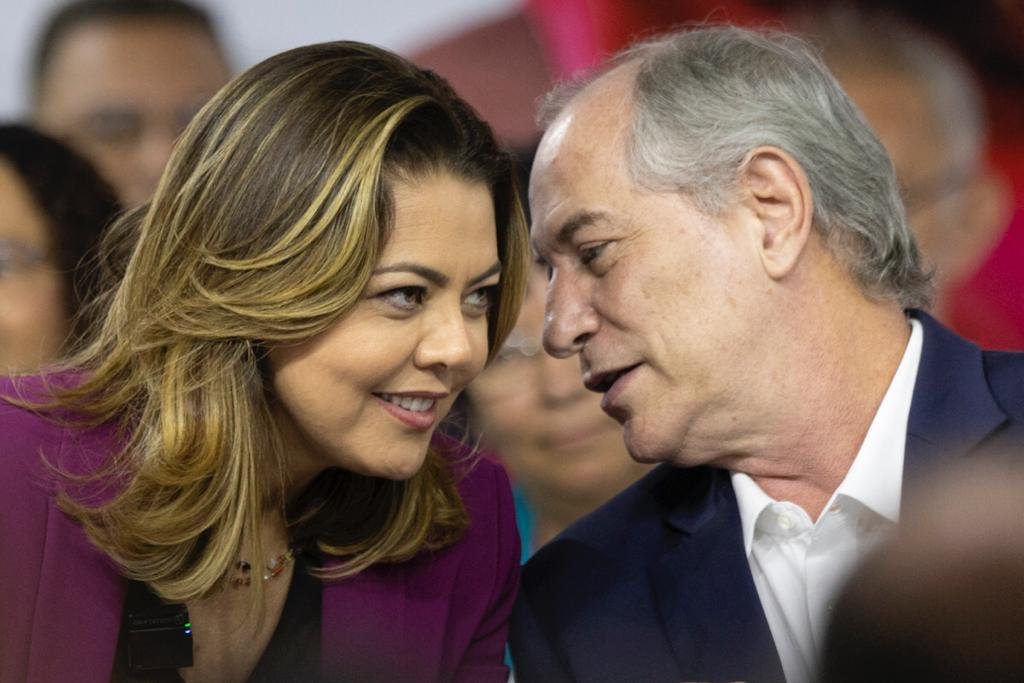 Mulher loira com a cabeça para o lado falando com homem. Ele é branco, careca e está de terno e também está com a cabeça para o lado