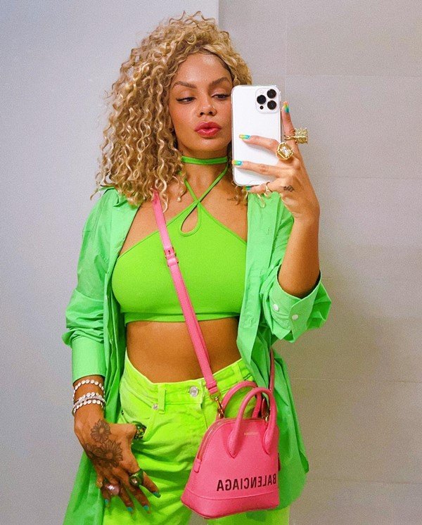 Mulher jovem e negra, com cabelo cacheado louro, posando para foto no espelho com um celular. Ela usa um top curto, uma camisa de botão, uma calça jeans, todos verdes, e uma bolsa rosa pink transpassada.