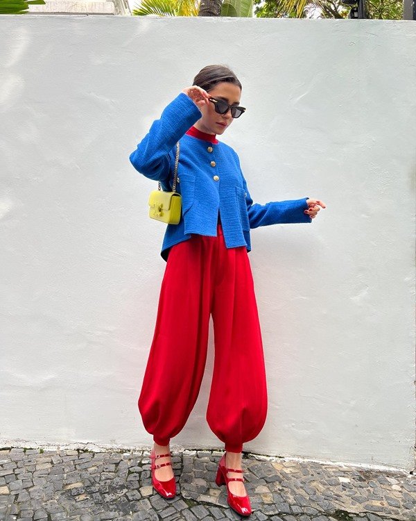 Mulher jovem e branca, com cabelo cacheado liso amarrado, posando para foto na rua, em um fundo branco. Ela usa um macacão com calças bufantes, em vermelho, e um casaco blazer azul forte por cima.