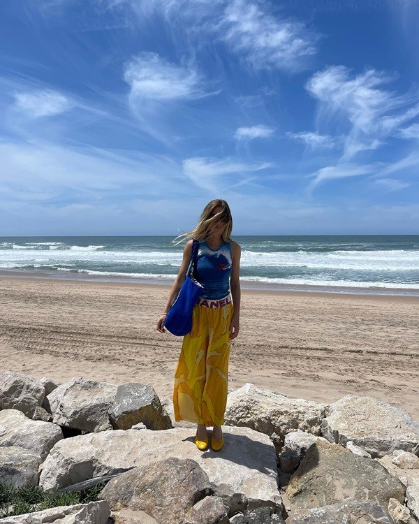 Mulher jovem e branca, com cabelo loiro longo, posando para foto na praia. Ela usa uma blusa azul, uma saia amarela e uma bolsa estilo sacola também azul.