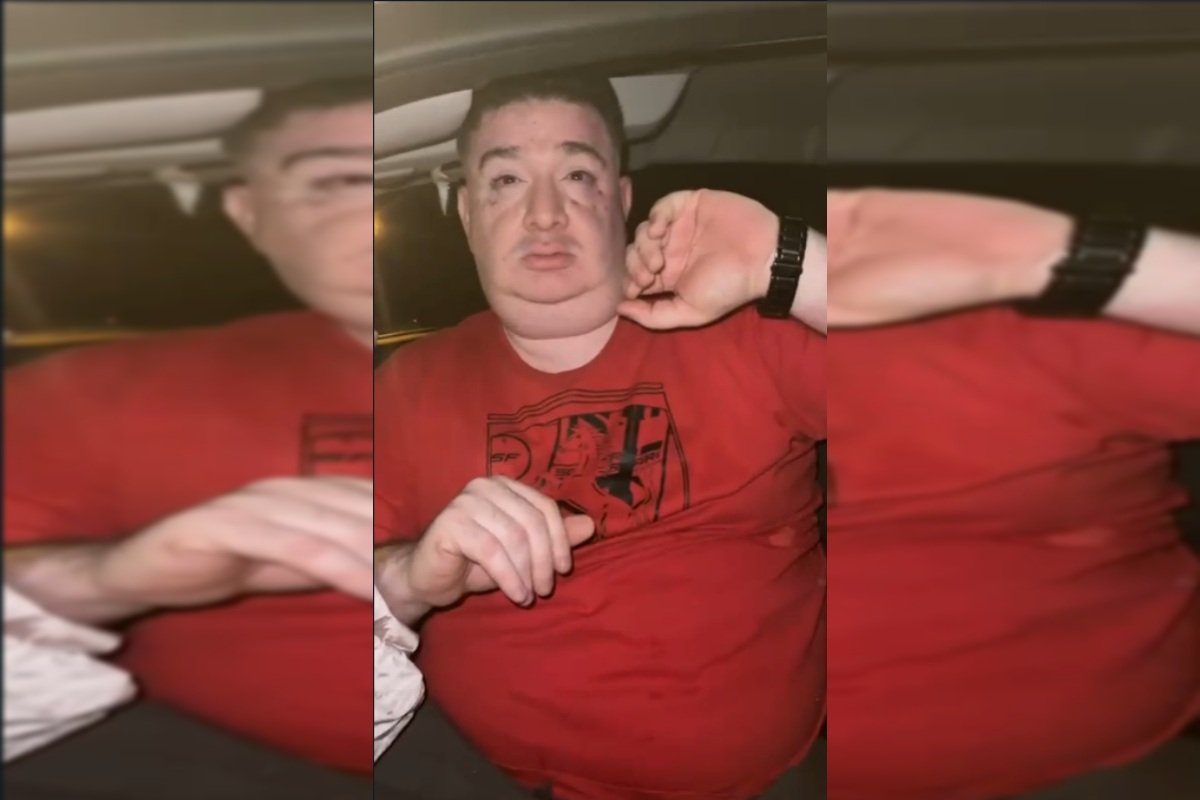 Homem branco de camisa vermelha coloca uma das mãos perto do rosto enquanto está sentado dentro de um carro