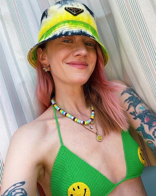 Mulher branca e jovem, de cabelos lisos e pintados de rosa, fazendo uma foto selfie. Ela usa um biquíni verde de crochê, colares de miçanga coloridos e um chapéu estilo pescador colorido 