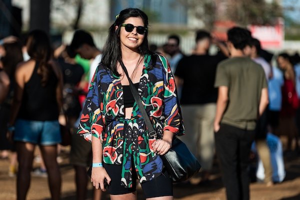 Mulher branca e jovem, de cabelo liso preto, posando para foto no Festival CoMA, em Brasília. Ela usa um top e um shorts preto e, por cima, um kimono colorido. Está de óculos escuros e uma bolsa preta transpassada.