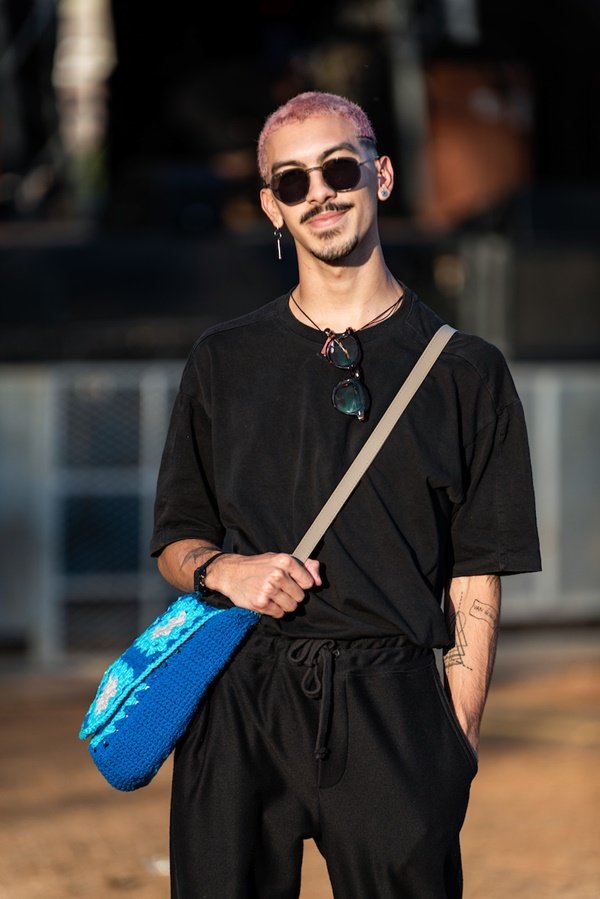 Homem jovem e negro, de cabelo curto descolorido, posando para foto no Festival CoMA, em Brasília. Ele usa um look todo preto: calça, camiseta e óculos escuros. Veste ainda uma bola de crochê azul transversal.