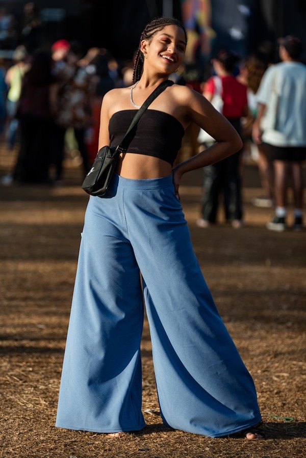 Mulher negra e jovem, de cabelo trançado longo, posando para foto no Festival CoMA, em Brasília. Ela usa um top preto sem alça, colar de prata, uma calça azul folgada e uma bolsa preta transpassada