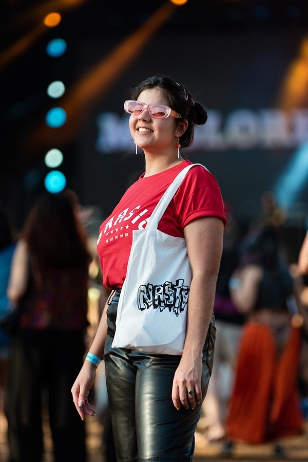 Mulher branca e jovem, de cabelo liso preto, posando para foto no Festival CoMA, em Brasília. Ela usa uma camiseta vermelha escrito BRASIL em branco, uma calça de couro preta, uma sacola estilo ecobag branca e óculos com lente rosa