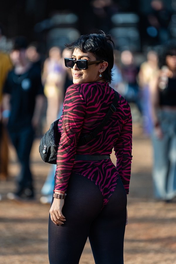 Mulher branca e jovem, de cabelo liso preto curto, posando para foto no Festival CoMA, em Brasília. Ela usa um maio de mangas cumpridas preto e rosa, meia calça preta, uma bolsa pochete preta e óculos escuros
