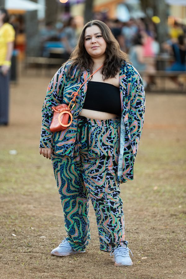 Mulher branca e jovem, de cabelo liso preto, posando para foto no Festival CoMA, em Brasília. Ela usa um top curto preto e um conjunto estampado de calça + kimono e uma bolsa transpassada laranja