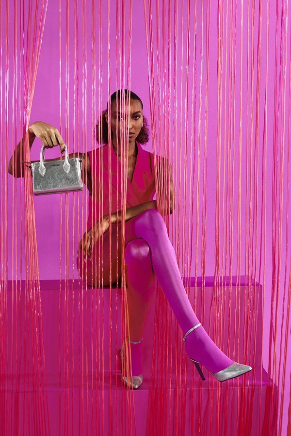 Young black woman with curly hair tied in a low ponytail posing for a photo in the Corello brand campaign.  She is on a pink background and wears a pink dress, pink tights, and heeled sandals and a totte bag, both silver.