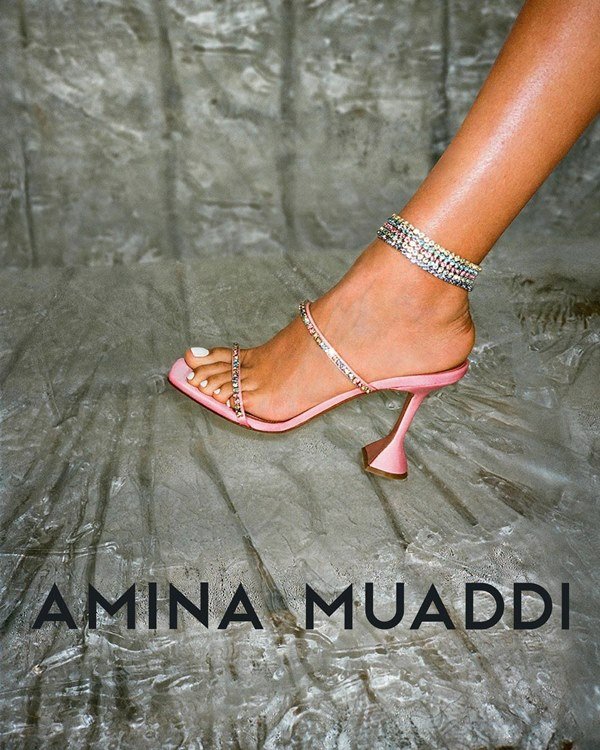 Campanha de divulgação da marca Amina Muaddi, onde é possível ver uma pessoa calçando uma sandália rosa de cetim com detalhes em estrasse