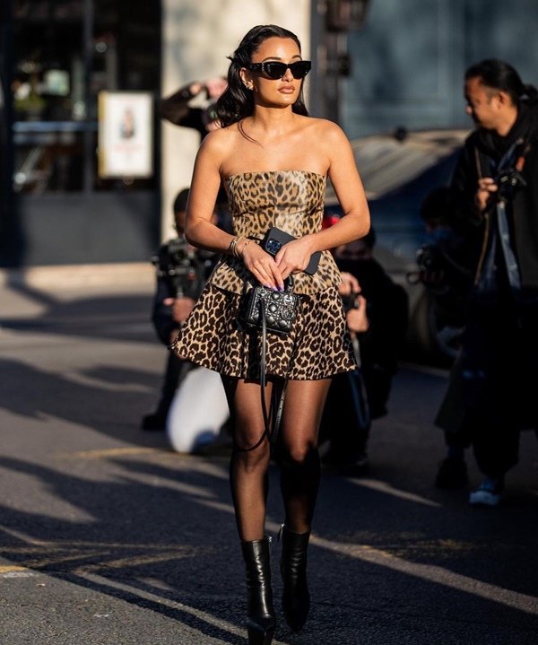 A estilista Amina Muaddi, uma mulher jovem de pele morena e cabelo liso longo, posando para foto na entrada do desfile da marca Dior. Ela usa um vestido curto e sem alças de onça, óculos escuros e botas pretas de couro
